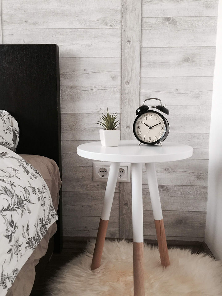 Ship small furniture. Cream bedside table with legs that are cream at the top and oak at the bottom. A small clock and cacti are perched on top of the table. 