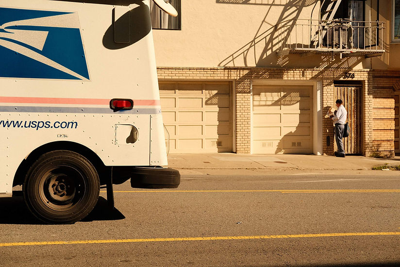 USPS truck in foreground with USPS guy in background delivering small package. This photo is to demonstrate USPS are cheaper for businesses delivering small packages vs. UPS. 